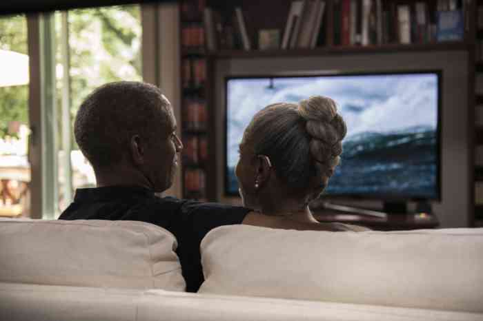 A man and a woman wearing hearing aids are sitting on a sofa watching tv