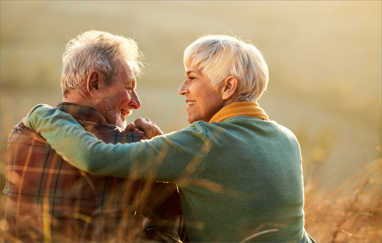 Couple de personnes âgées souriant tendrement l’un à l’autre, enlacés dans un paysage naturel au coucher du soleil.