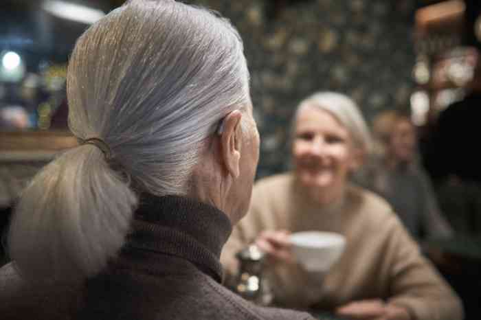 two elderly friends drinking tea, one wearing a hearing aid.