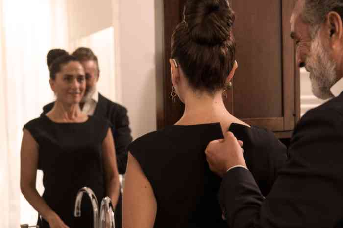 Couple dans la salle de bain se préparant à sortir : l'homme ferme la robe de sa femme, qui porte un appareil auditif.