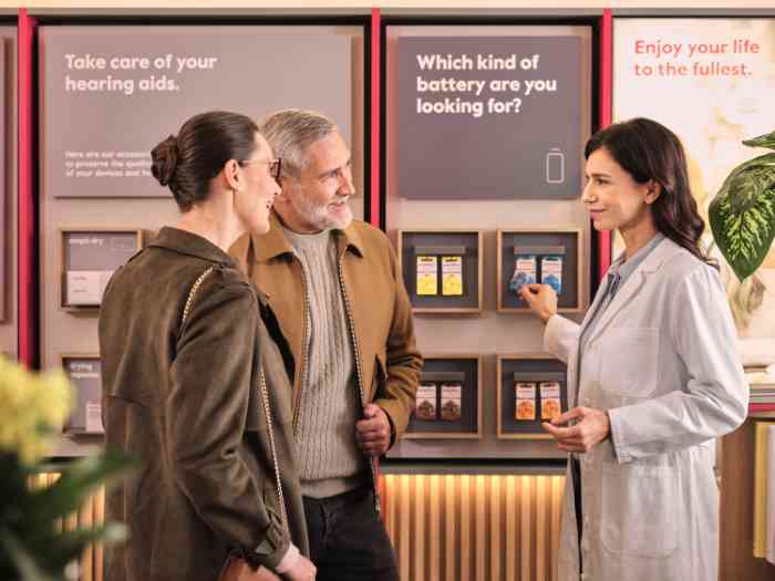 Ein Paar und ein Audiologe im Kittel sprechen vor einem Panel, das Hörgerätebatterien ausstellt, englisches Geschäft.