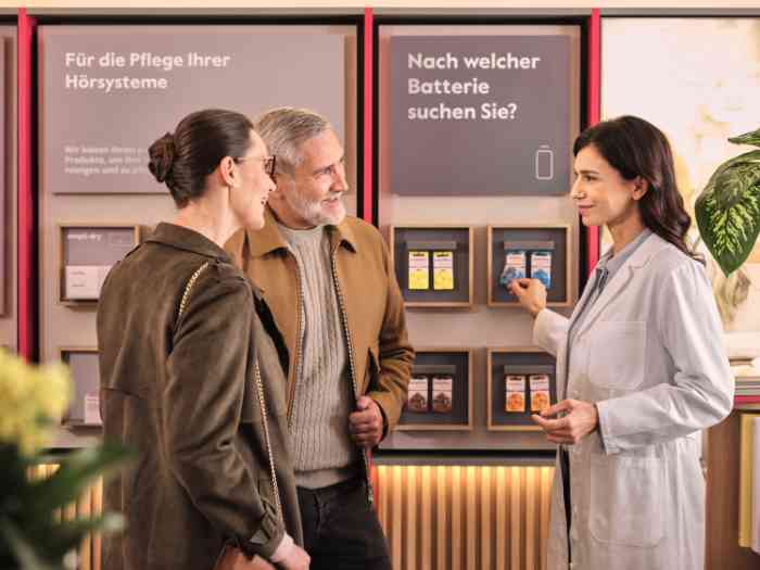 Ein Paar und ein Audiologe im Kittel sprechen vor einem Panel, das Hörgerätebatterien ausstellt, deutsches Geschäft.