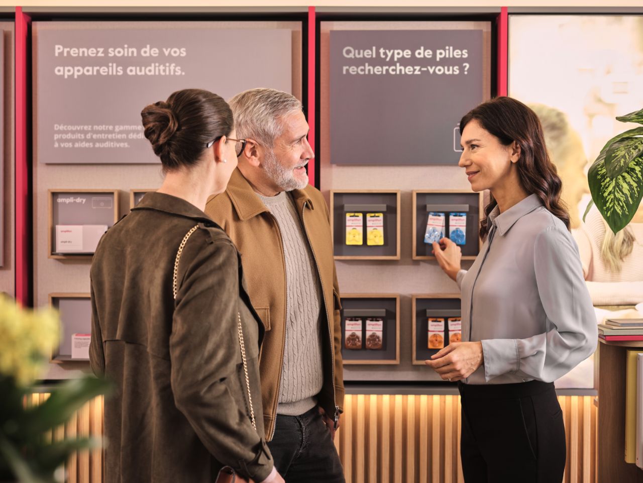 Couple et audiologiste sans blouse devant un panneau de piles pour appareils auditifs, magasin français.