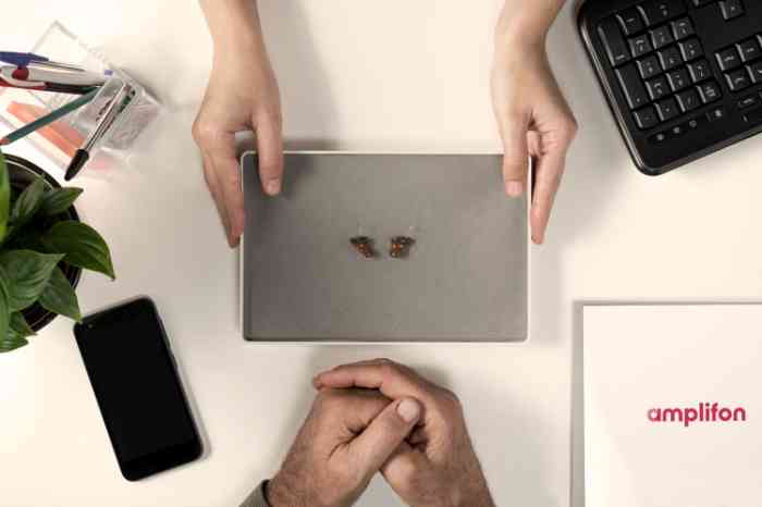 Two hearing aids on gray tray, hands pointing towards them.