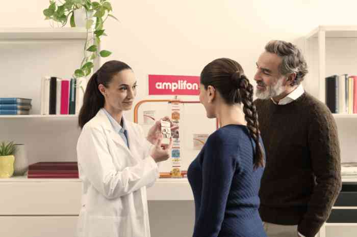 Audiologist in lab coat showing Amplifon hearing aid batteries to two clients in store.
