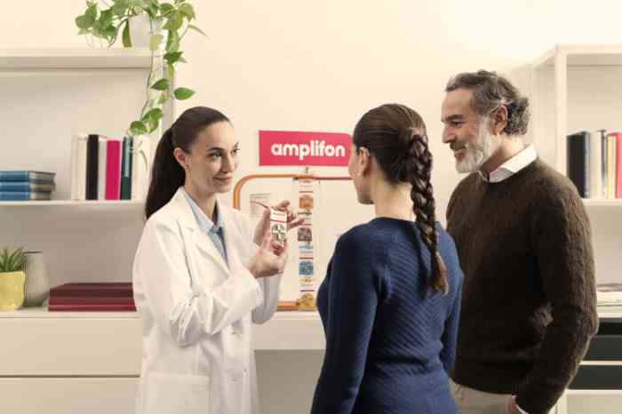 Audiologist in lab coat showing Amplifon hearing aid batteries to two clients in store.