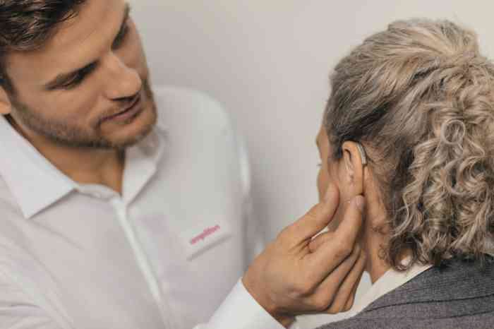 Male audiologist adjusting hearing aid on elderly female client's ear.