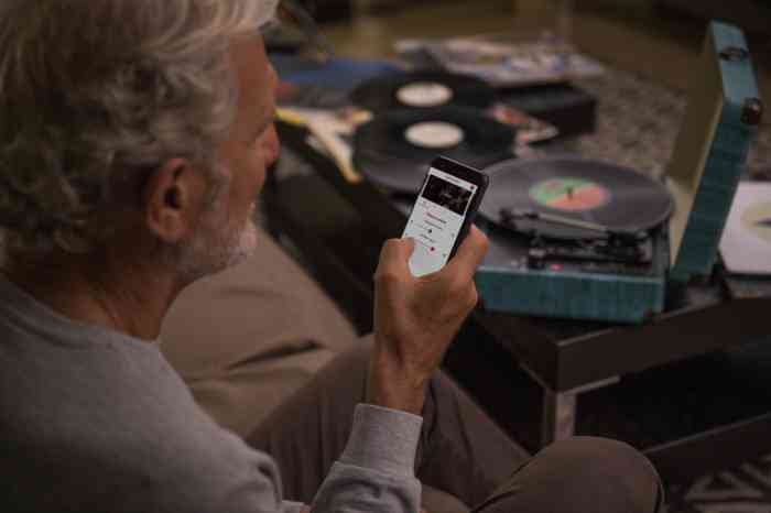 Un homme âgé assis sur un canapé consulte son téléphone portable en écoutant des disques vinyls.