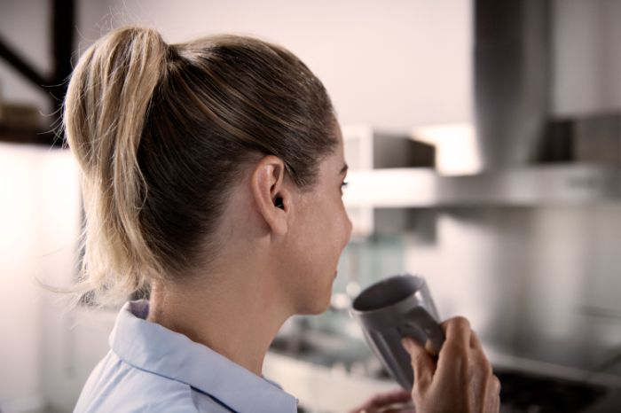 Une jeune femme de profil dans sa cuisine en train de boire, un mug à la main.