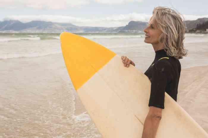 Femme senior vue de profil sur une plage face à la mer, tenant une planche de surf.