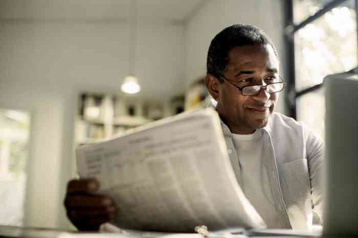 A man with glasses working at his computer and holding a newspaper