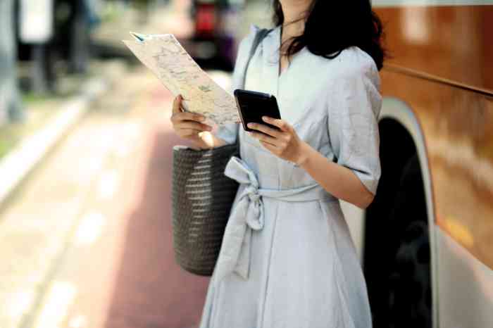 Femme en robe bleue devant un autocar tenant une carte et une tablette dans ses mains.