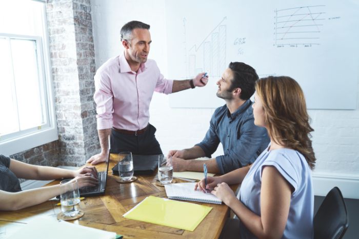 4 personnes en réunion d'entreprise : 3 personnes sont assises à une table prenant des notes et un homme se tient debout en pointant du doigt un tableau au mur.