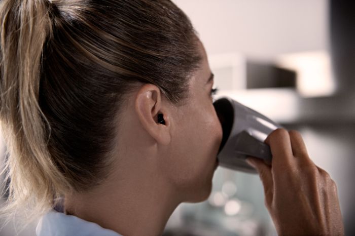 Femme vue de profil portant une aide auditive à l'intérieur de son oreille gauche et buvant dans un mug.